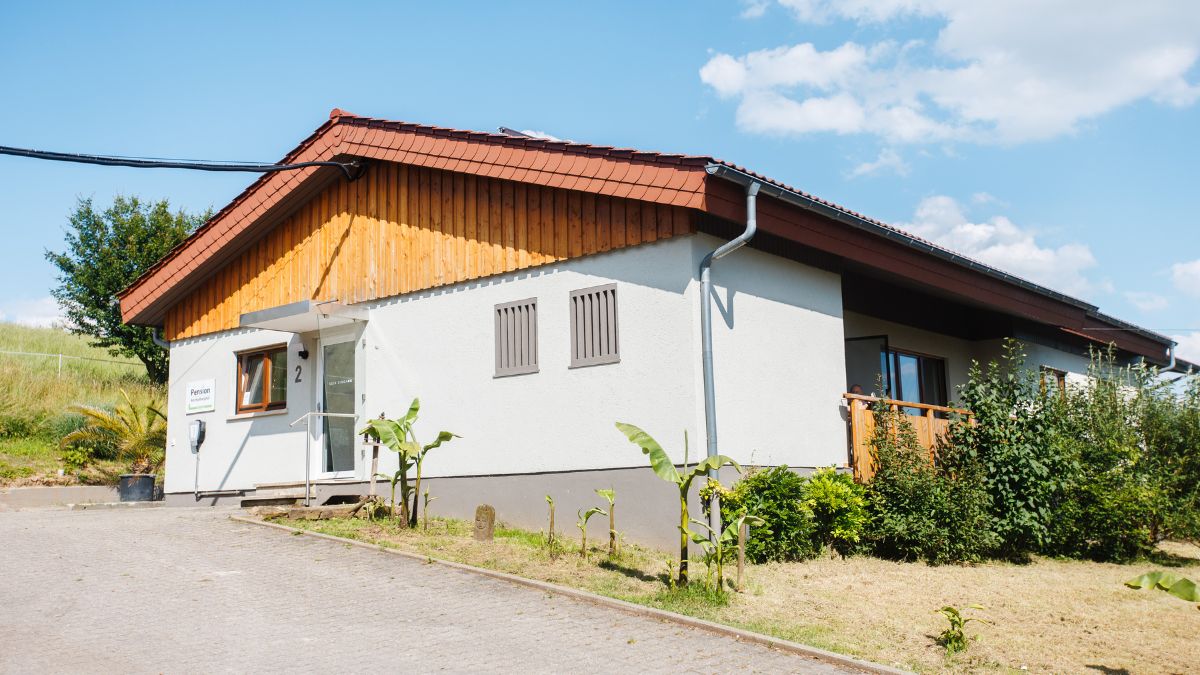Weißes Gebäude mit rotem Ziegeldach und Holzverkleidung am Giebel, Eingangsbereich der Pension auf dem Hof