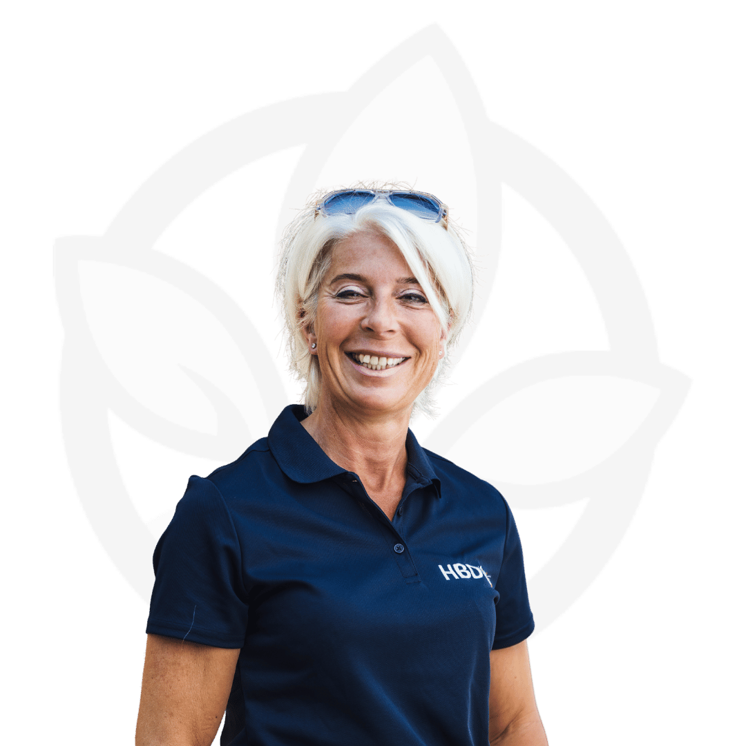 Lächelnde Anja Beifuss von HBD Vital® in dunkelblauem Polo-Shirt mit HBD-Logo, Sonnenbrille im Haar, vor hellem Hintergrund mit Blattmotiv