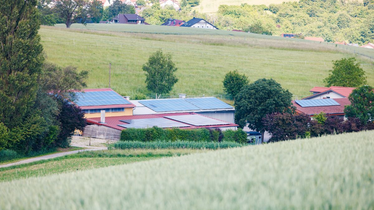 Blick von einem Hügel über Felder auf den Hohberghof mit mehreren Gebäuden, deren Dächer mit Solaranlagen bestückt sind, umgeben von Wiesen und Bäumen